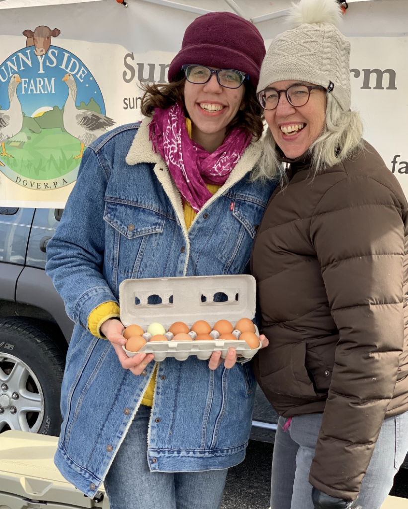 Two farmers, a mother and a daughter, wearing winter hats and holding a carton of farm-fresh eggs